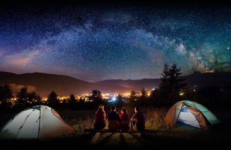 Four friends sitting on a bench between two lit tents under the Milky Way at a hillside campsite